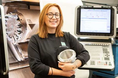 Mitarbeiterin mit Werkst&uuml;ck vor der CNC-Drehmaschine